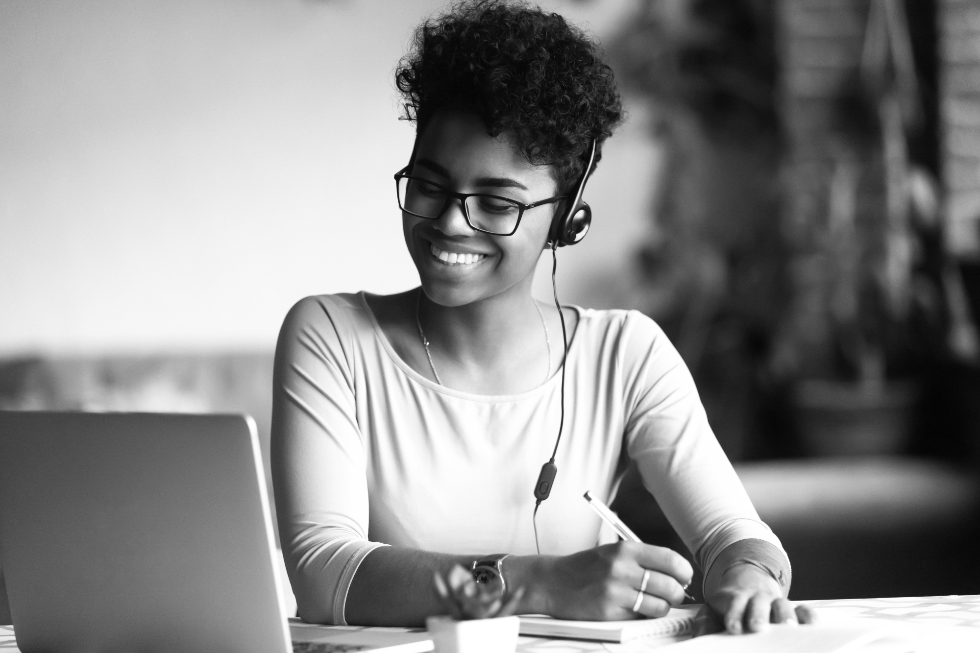 Grayscale photo of a young student of color wearing glasses, headphones, and smiling at a laptop while taking notes.
