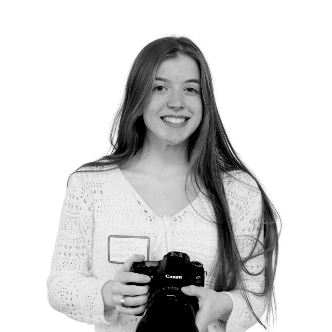Grayscale photo of Aspirations in Computing (AiC) Campus Rep Sarah Olson smiling toward the viewer and holding a DSLR camera 